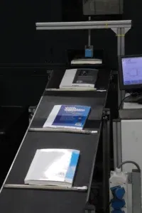 Boeken op een transportband in een industriële omgeving, tijdens een geautomatiseerd productieproces met een machine en beeldscherm op de achtergrond.