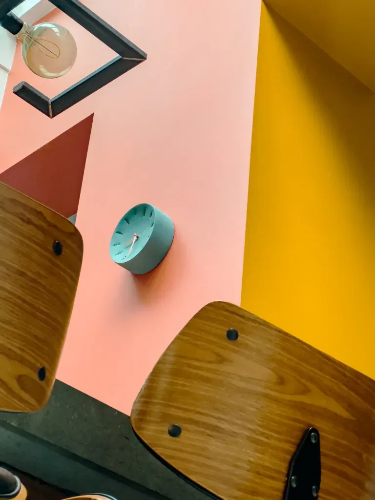 Modern interieur met een roze en gele muur, een blauwgroene klok, houten stoelen en een geometrische lamp met een zichtbare gloeilamp.
